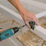 Unrecognizable professional male master using cordless screwdriver to install metal hinge on wooden window in light room during repair works